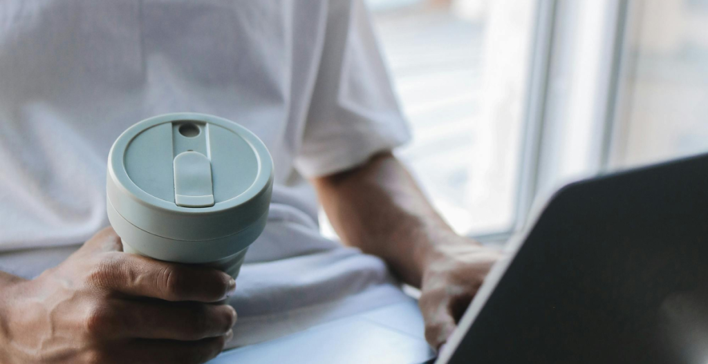 Person holding a reusable travel coffee mug while working on a laptop near a window.