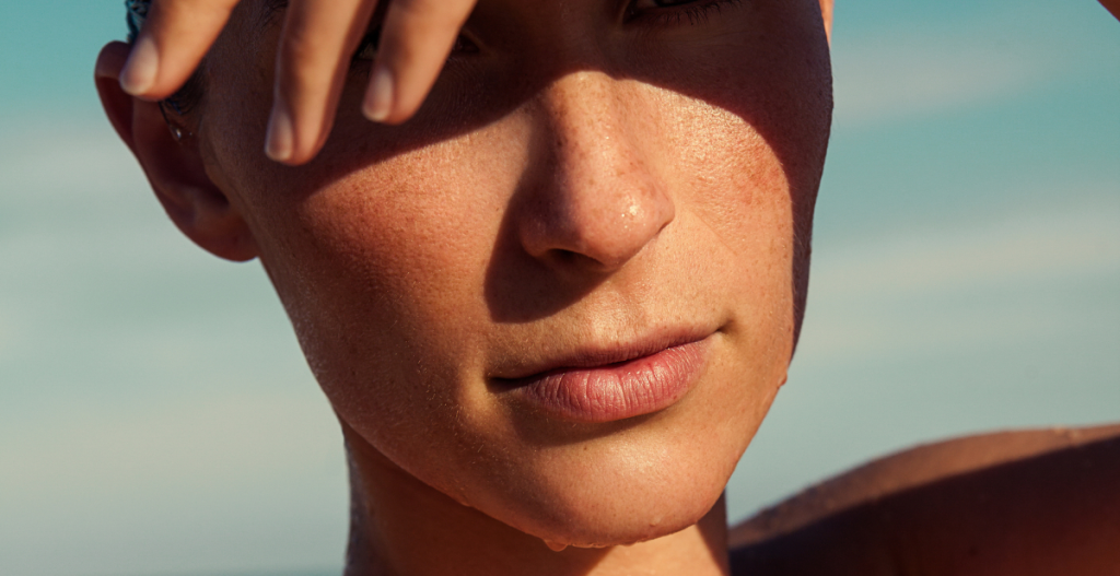 Close-up of a person shielding their face from the sun with their hand, showing sun-kissed skin with visible freckles and texture.